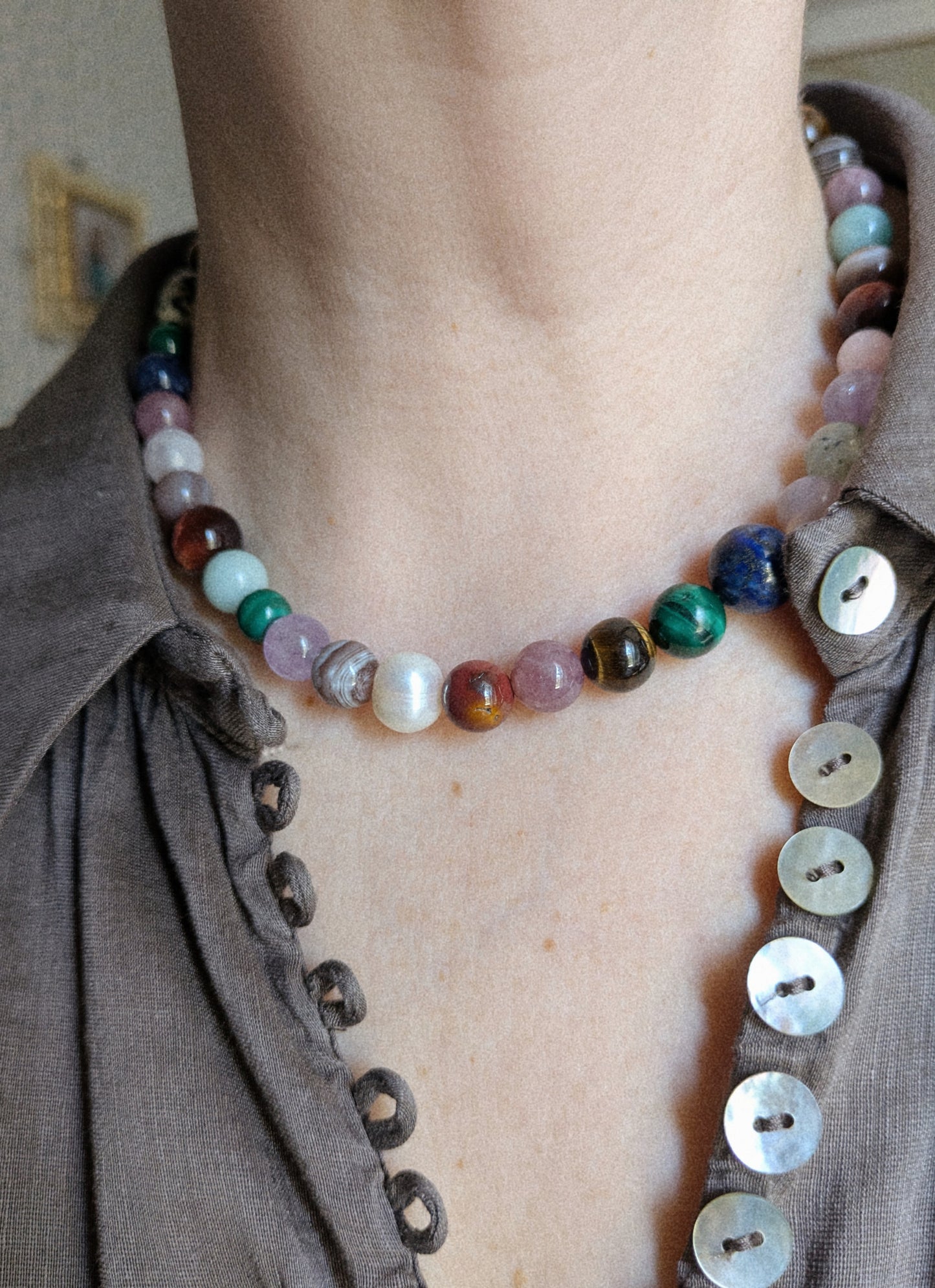 A close-up of a person's neck and chest wearing a beaded choker necklace made of polished multicolored gemstone-like beads, paired with a brown button-front shirt.