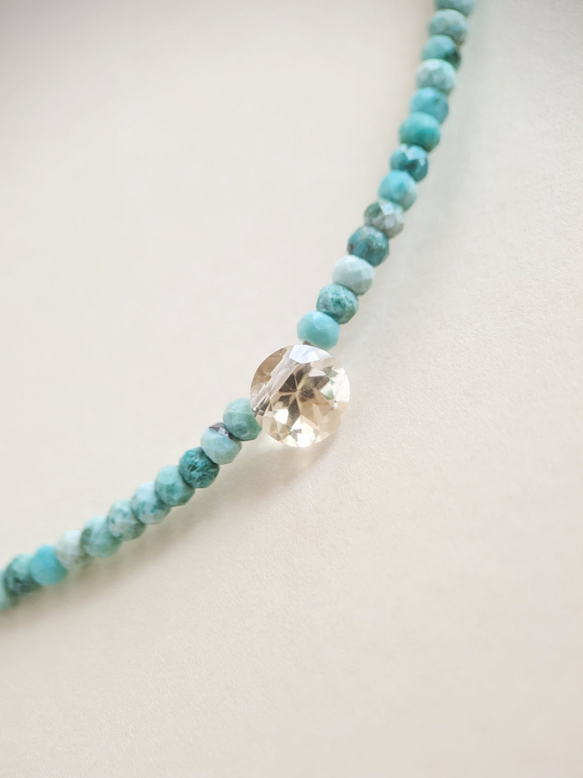 A beaded necklace featuring light blue and turquoise marbled beads with a central faceted citrine bead, displayed on a neutral cream background.
