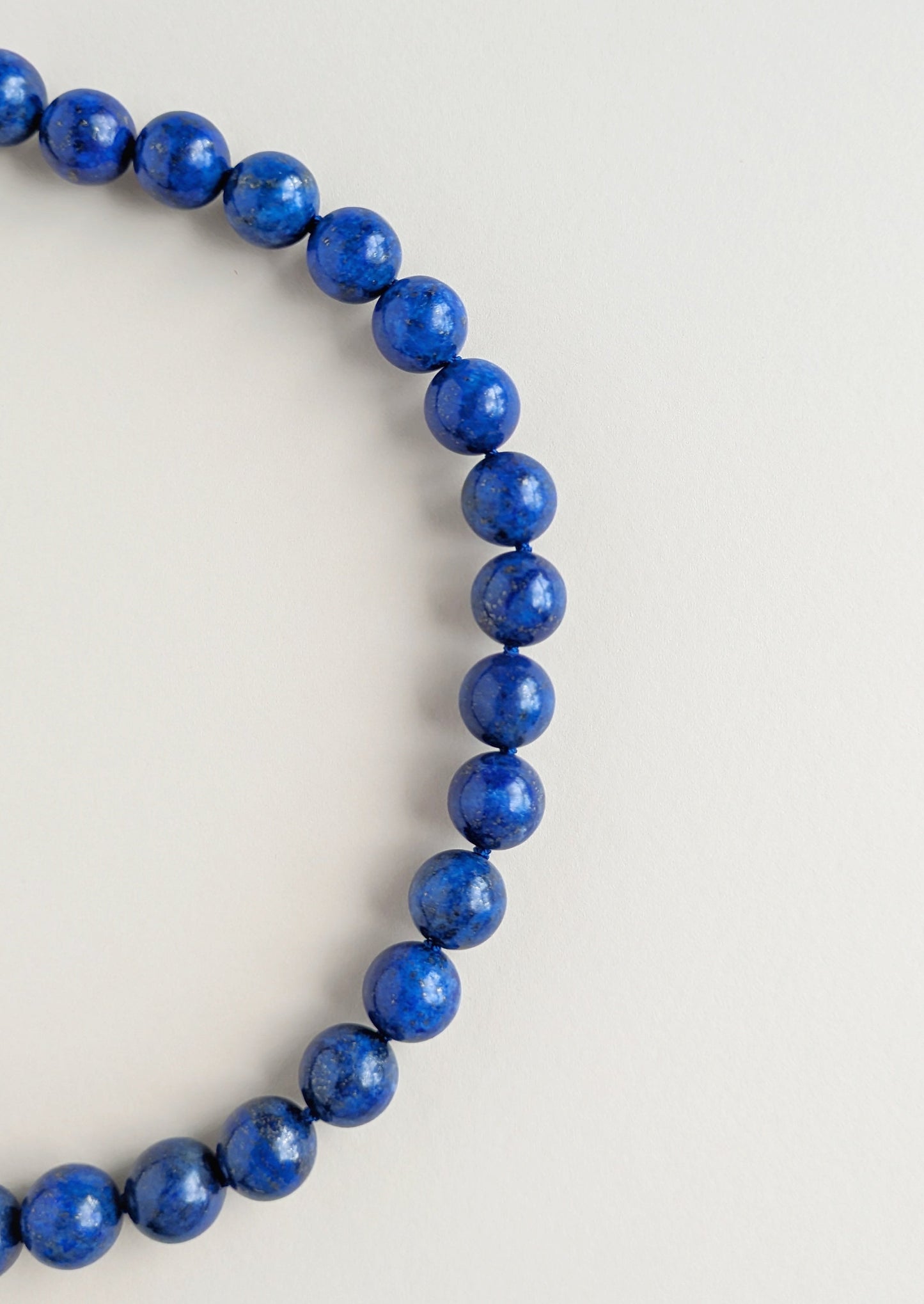 A strand of polished blue beads,lapis lazuli, arranged in a curved circle on a plain light gray background.