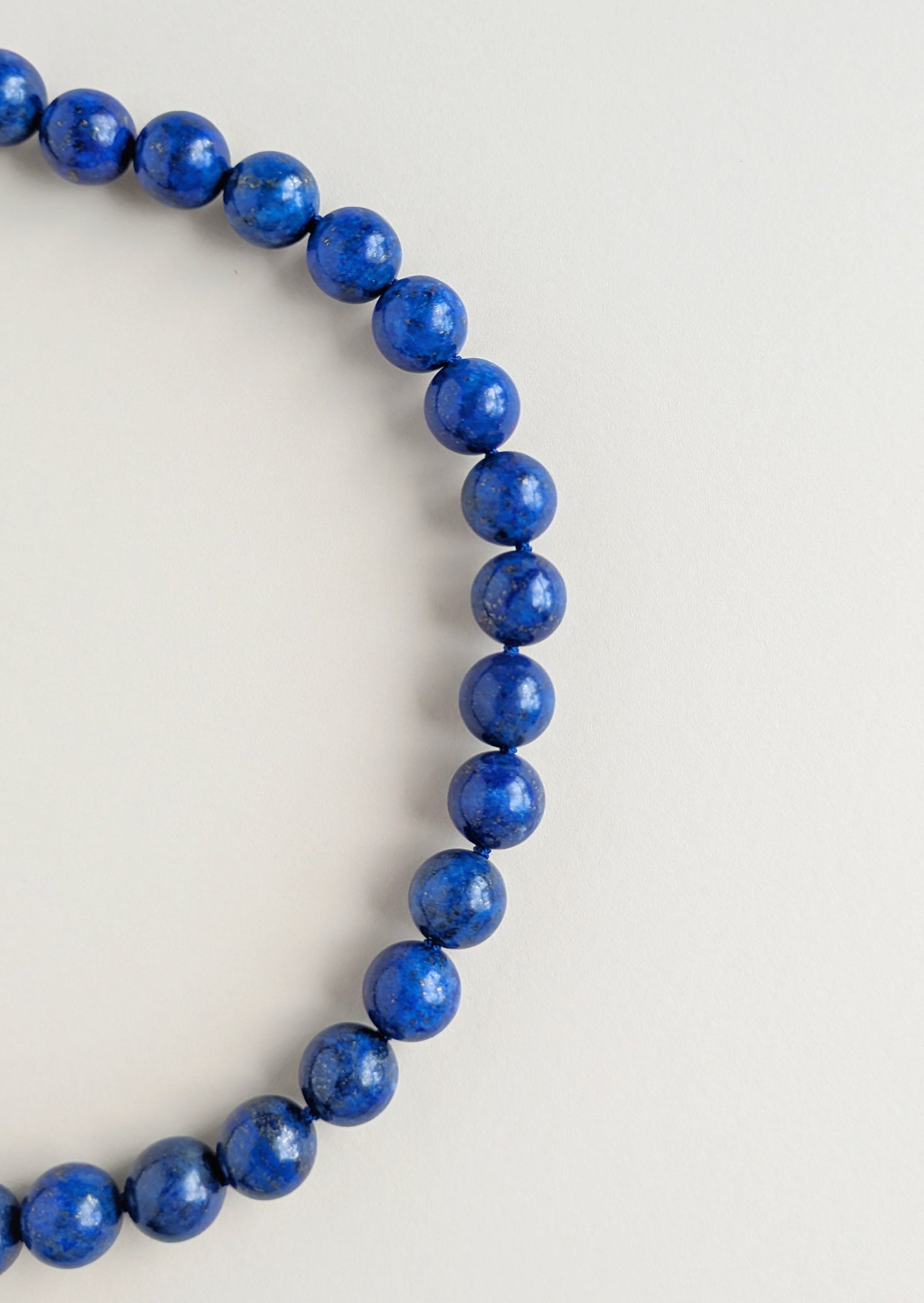 A strand of polished blue beads,lapis lazuli, arranged in a curved circle on a plain light gray background.