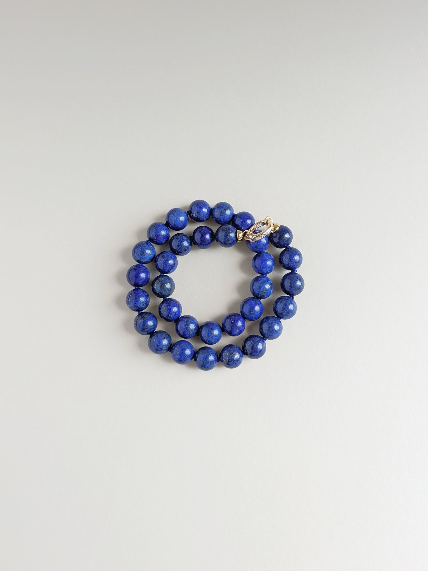 A wrapped around necklace made of polished royal blue lapis lazuli stones, featuring a gold-toned toggle clasp, displayed on a neutral light gray background.