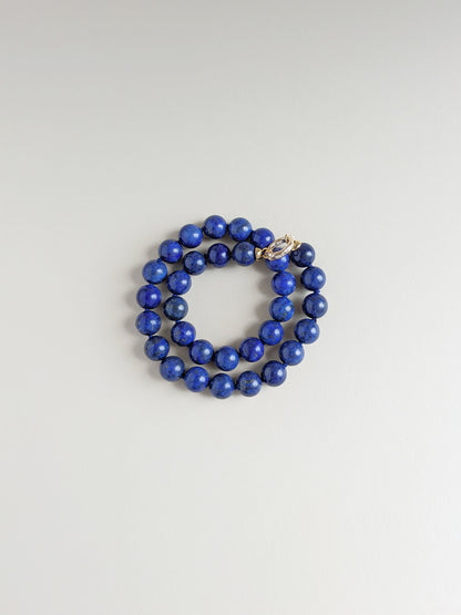 A wrapped around necklace made of polished royal blue lapis lazuli stones, featuring a gold-toned toggle clasp, displayed on a neutral light gray background.
