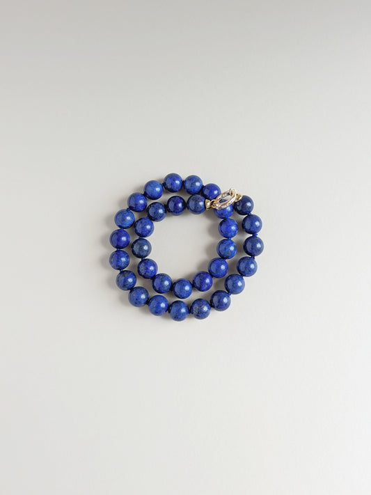 A wrapped around necklace made of polished royal blue lapis lazuli stones, featuring a gold-toned toggle clasp, displayed on a neutral light gray background.