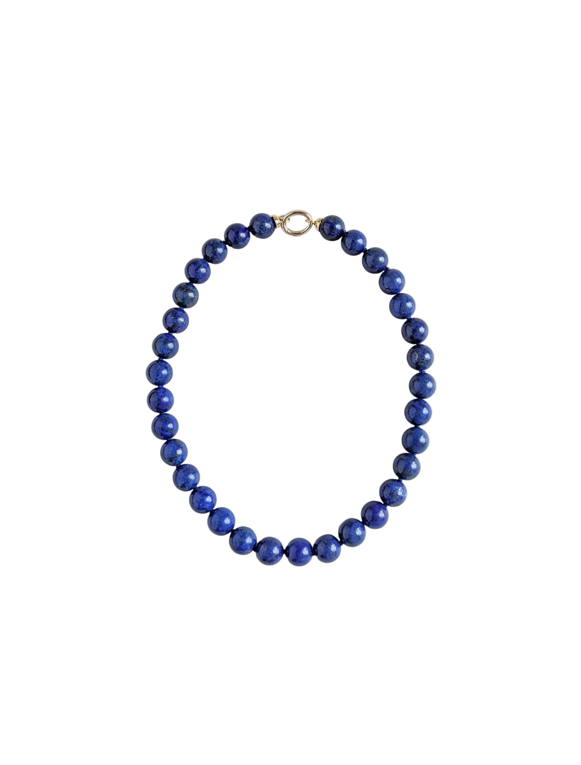 A beaded necklace made of round, polished dark blue lapis lazuli stones, arranged in an oval shape with a small gold clasp at the top, set against a plain light gray background.
