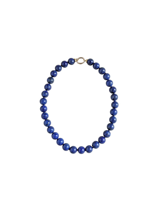A beaded necklace made of round, polished dark blue lapis lazuli stones, arranged in an oval shape with a small gold clasp at the top, set against a plain light gray background.