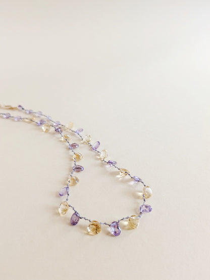 A delicate beaded necklace featuring faceted amethyst and citrine teardrop-shaped gemstones
 on a thin silver wire, laid flat on a warm beige background.