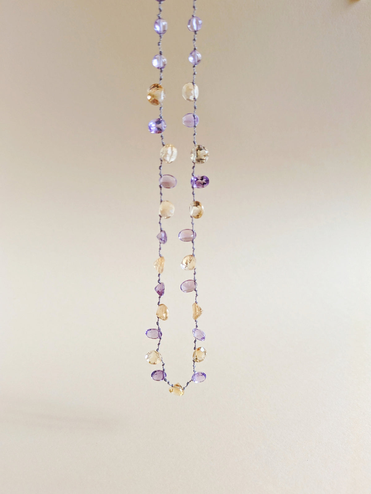 A delicate beaded necklace featuring faceted amethyst and citrine teardrop-shaped gemstones
 on a thin silver wire, laid flat on a warm beige background.