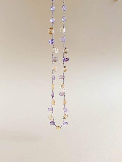 A delicate beaded necklace featuring faceted amethyst and citrine teardrop-shaped gemstones
 on a thin silver wire, laid flat on a warm beige background.