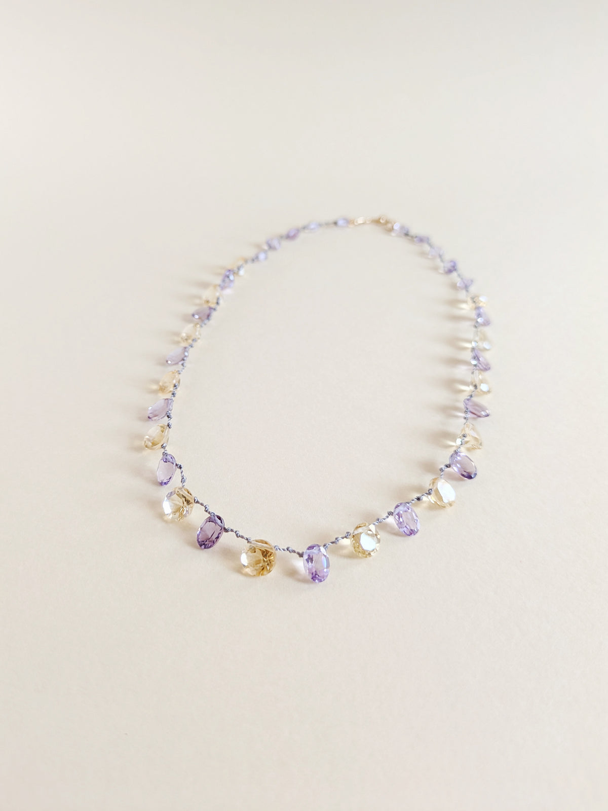 A delicate beaded necklace featuring faceted amethyst and citrine teardrop-shaped gemstones
 on a thin silver wire, laid flat on a warm beige background.