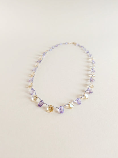 A delicate beaded necklace featuring faceted amethyst and citrine teardrop-shaped gemstones
 on a thin silver wire, laid flat on a warm beige background.