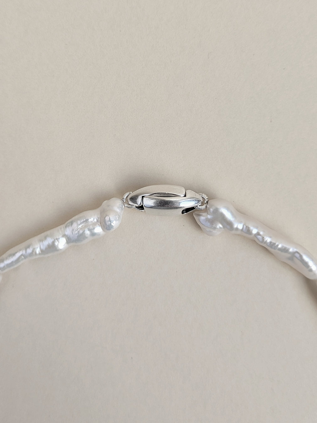 A close-up of a silver clasp of pearl choker made of irregular keshi biwa pearls on a plain beige background.