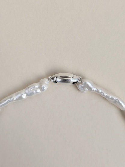 A close-up of a silver clasp of pearl choker made of irregular keshi biwa pearls on a plain beige background.
