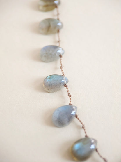 A close-up of a necklace featuring teardrop-shaped labradorite stones strung on a thin brown cord, set against a solid light beige background.