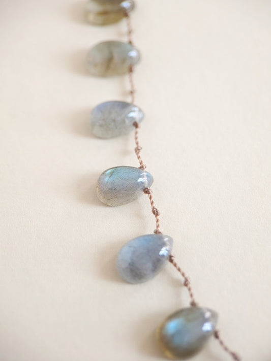 A close-up of a necklace featuring teardrop-shaped labradorite stones strung on a thin brown cord, set against a solid light beige background.