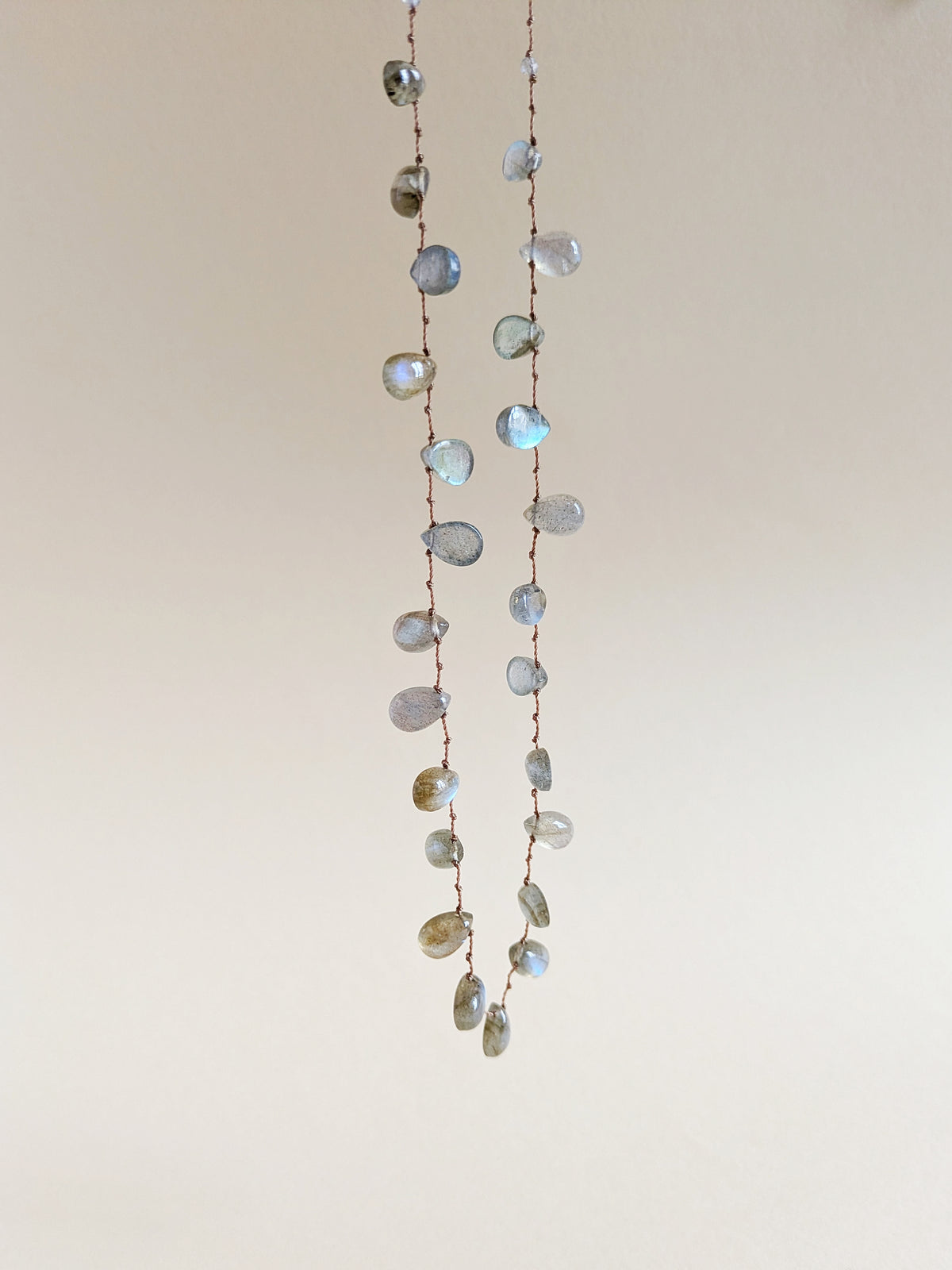 A long strand necklace made of labradorite teardrop beads on a thin brown cord, hanging against a plain light beige wall.