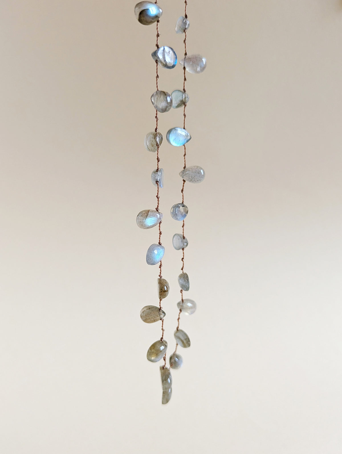 A long strand necklace made of labradorite teardrop beads on a thin brown cord, hanging against a plain light beige wall.