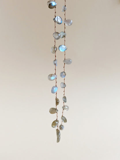 A long strand necklace made of labradorite teardrop beads on a thin brown cord, hanging against a plain light beige wall.