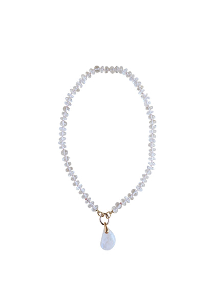 A delicate beaded necklace with clear faceted beads and a small white teardrop moonstone pendant with gold-tone metal accents, displayed on a plain light beige background.