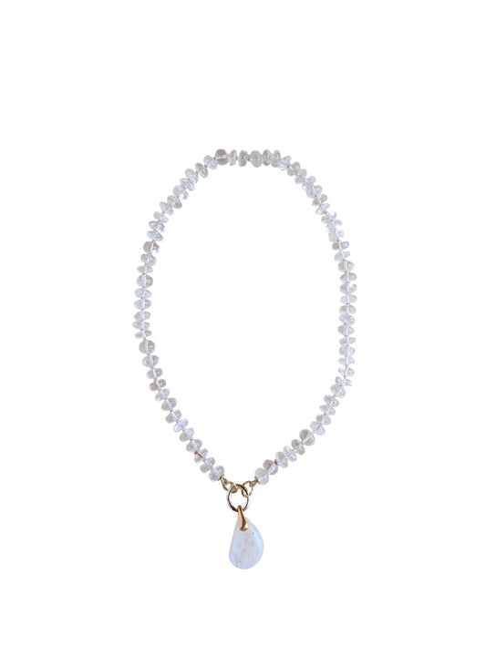 A delicate beaded necklace with clear faceted beads and a small white teardrop moonstone pendant with gold-tone metal accents, displayed on a plain light beige background.
