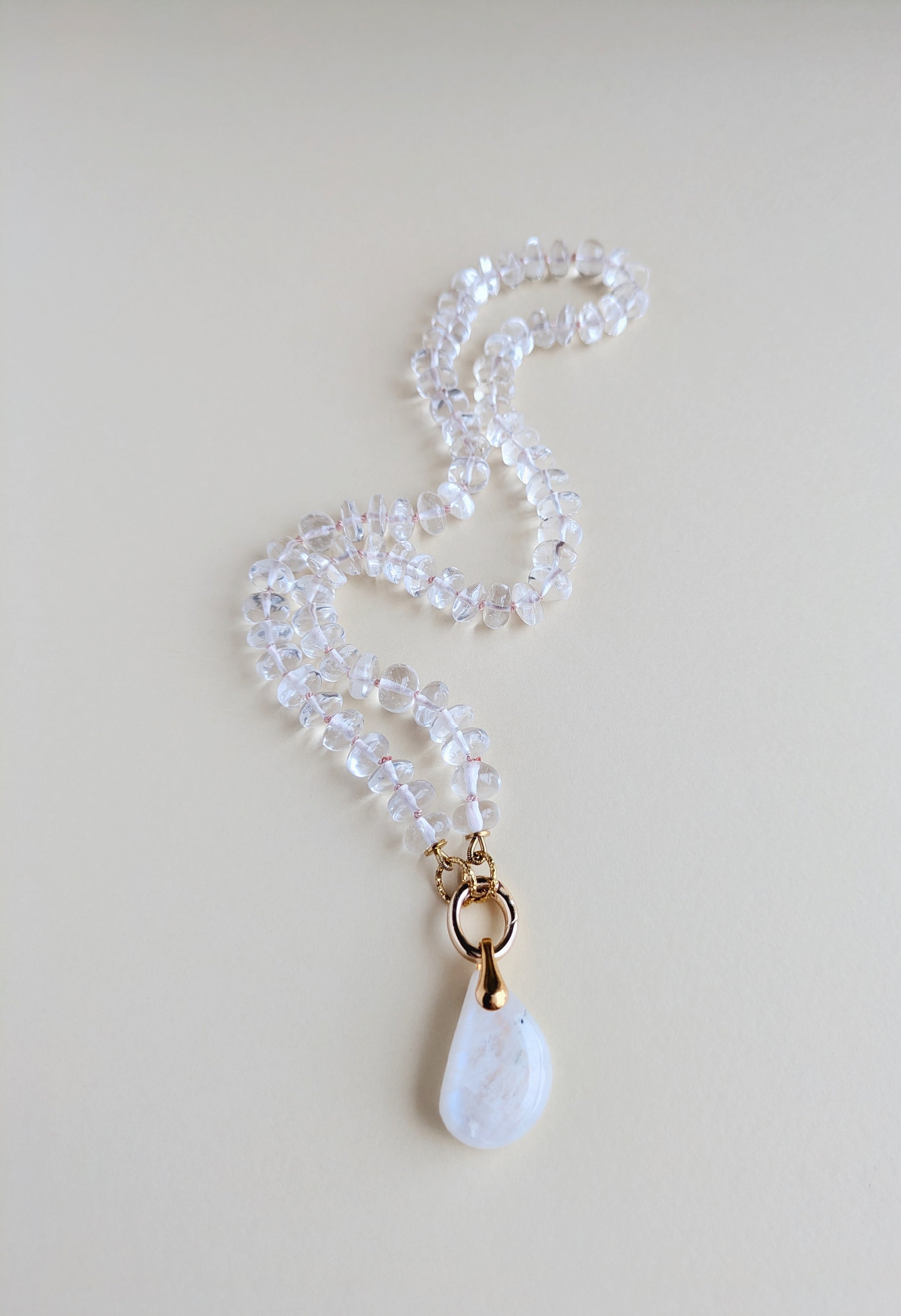 A beaded necklace made of clear faceted beads with a gold-tone ring and a white teardrop-shaped moonstone pendant, displayed on a light beige background.