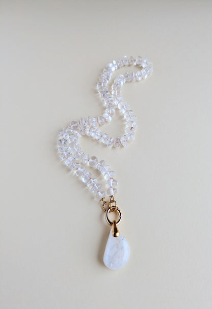A beaded necklace made of clear faceted beads with a gold-tone ring and a white teardrop-shaped moonstone pendant, displayed on a light beige background.