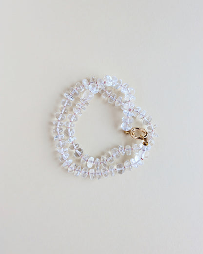 A delicate beaded bracelet made of clear faceted beads with a small gold clasp, photographed on a plain light beige background.