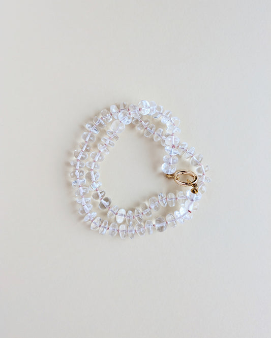 A delicate beaded bracelet made of clear faceted beads with a small gold clasp, photographed on a plain light beige background.