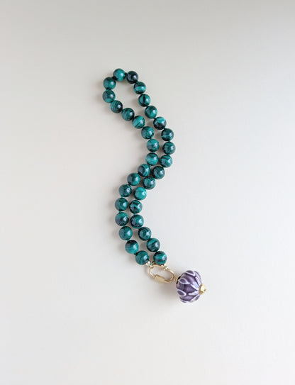 A beaded necklace with glossy malachite beads and a purple patterned pendant, displayed on a clean white background.