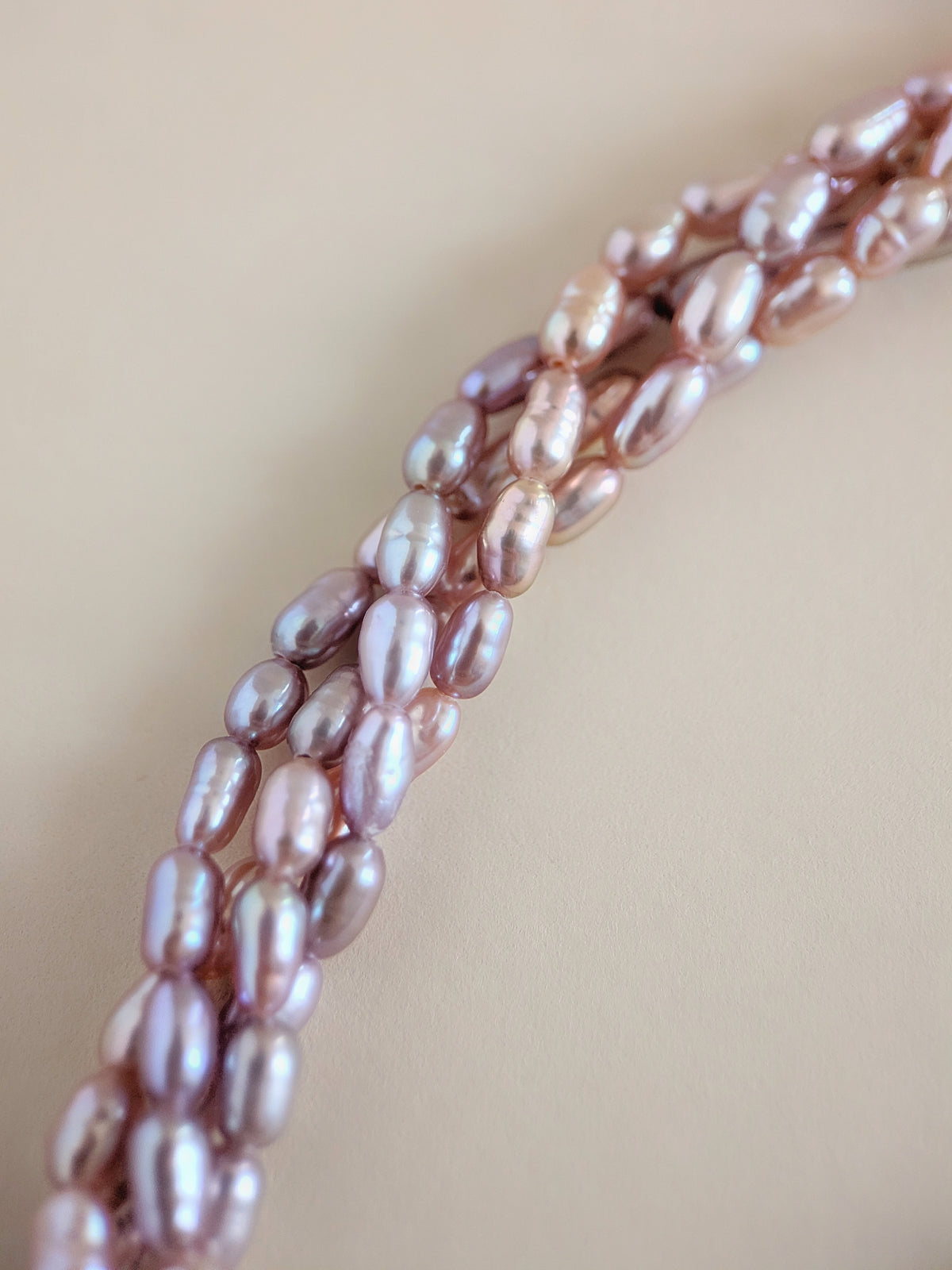 Close-up of a strand of irregular baroque pearls in a soft pink-lilac tone, arranged diagonally on a warm beige background.