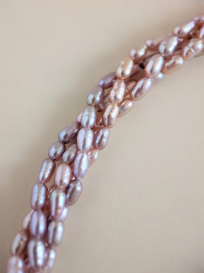 Close-up of a strand of irregular baroque pearls in a soft pink-lilac tone, arranged diagonally on a warm beige background.