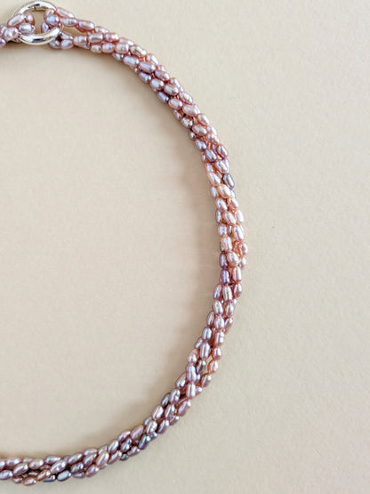 A close-up of a beaded chain necklace with small, irregular pearlescent beads in a metallic rose gold and silver tone, arranged in a loop on a solid beige background.