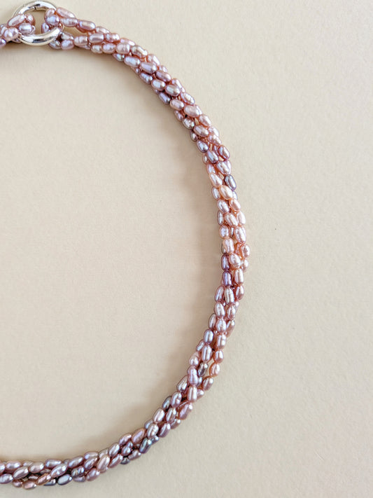 A close-up of a beaded chain necklace with small, irregular pearlescent beads in a metallic rose gold and silver tone, arranged in a loop on a solid beige background.